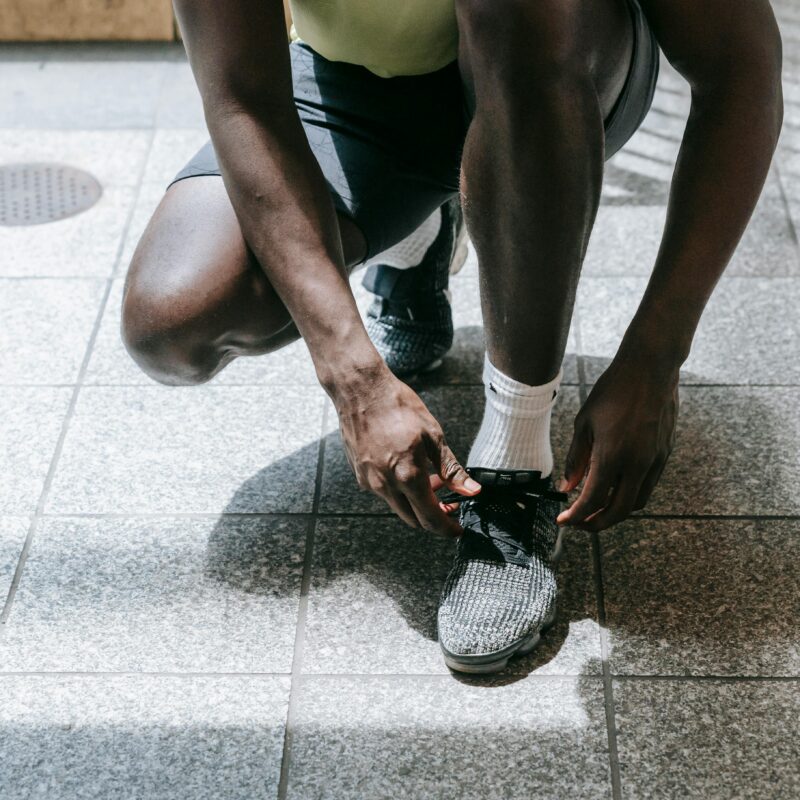 Person kneeling on a tiled floor, tying the laces of a gray athletic shoe with white socks nearby